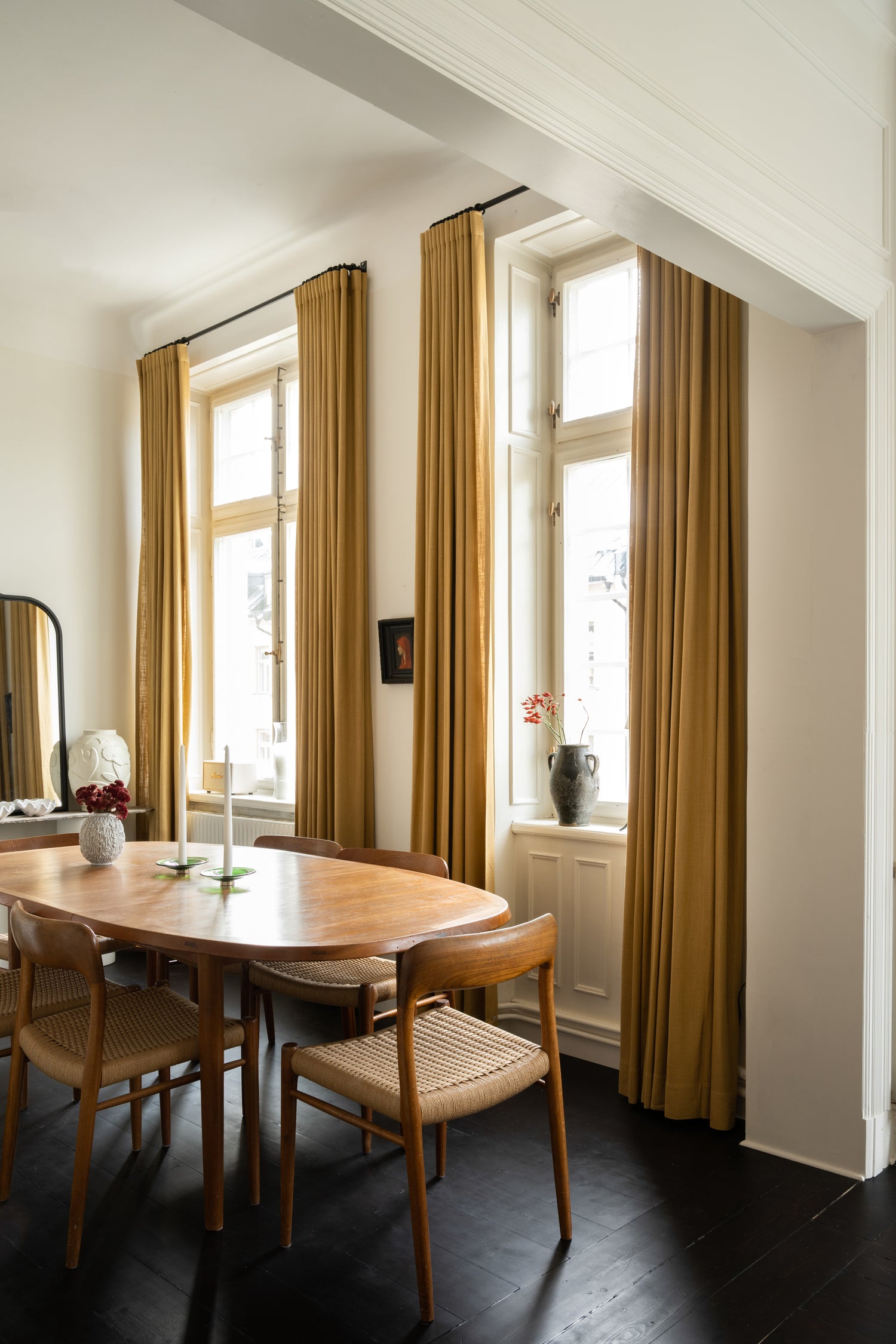 Bilde av en klassisk leilighet, stue med hvite vegger, malte svarte gulv. Sennepsgule okergule gardiner i svart gardinstang. Retro møbler: Ovalt spisebord og spisestoler i varmt treverk. 