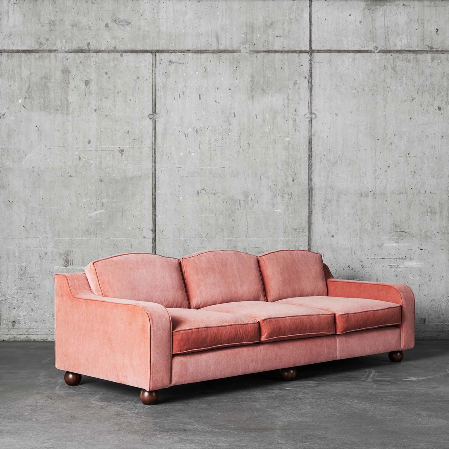 Bilde av den rosa sofaen Lola i fargen Velvet Vintage Pink fra Dusty Deco mot rå betongvegger. Sofaen har mørkebrune ben. 