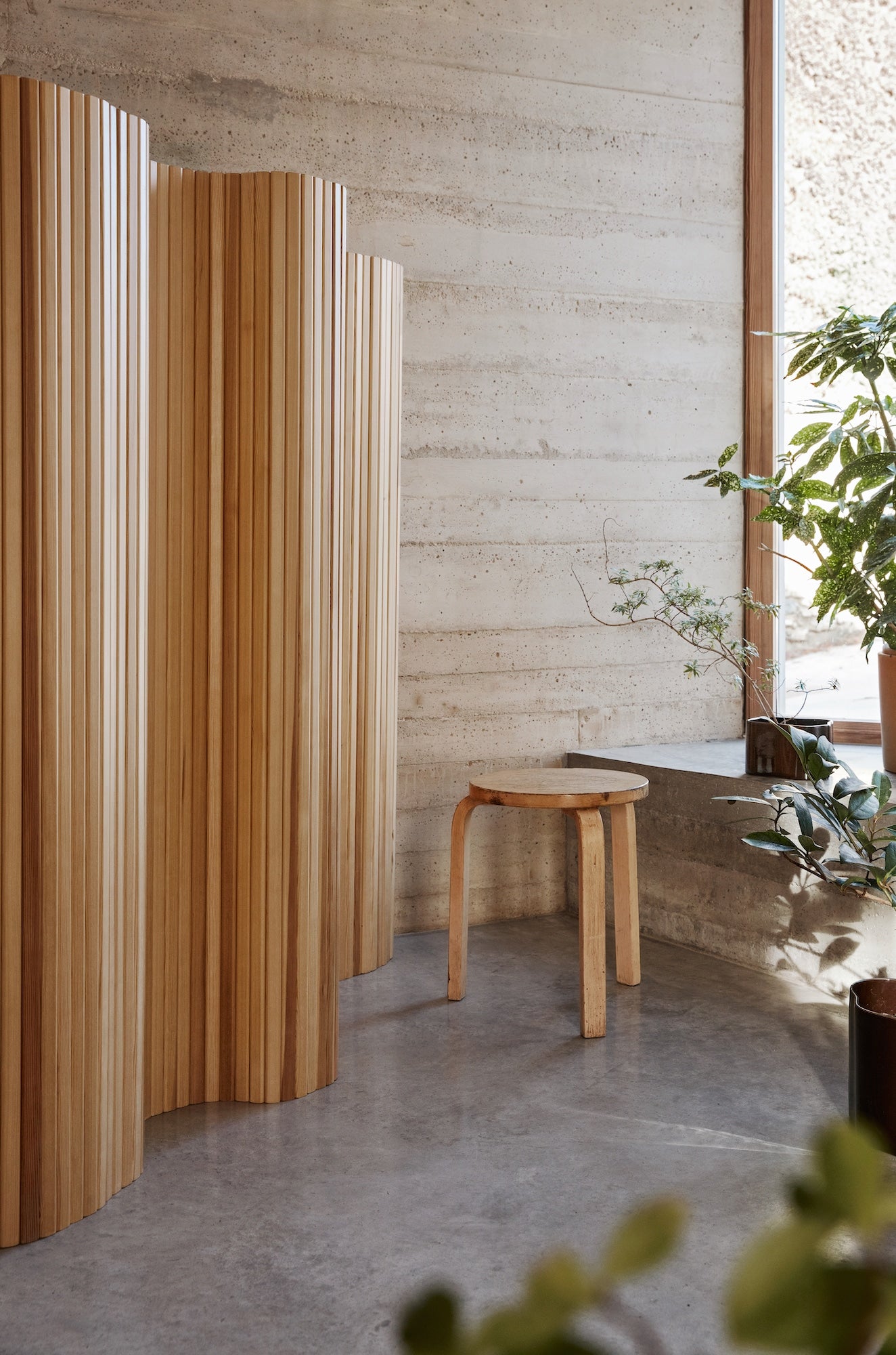 Bilde av skilleveggen Screen 100 fra Artek, designet av Alvar Aalto i 1936. Skjermen står i et rom med vegger og gulv av stein, grønne planter og krakken Stool 60, også fra Artek.