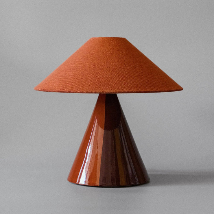 Bordlampen Caterina Cognac har en håndmalt tofarget lampefot i dyp mørkerød og konjakkfarget glasert keramikk og skjerm i konjakkfarget bomullsstoff som sprer et behagelig og stemningsfullt varmt lys. En elegant bordlampe i en helt unik kombinasjon av farger og teksturer!