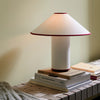 Den elegante bordlampen Colette ATD6 i Art Deco-stil fra &tradition har en spennende avantgarde-estetikk som får hjertet vårt til å slå litt fortere. Bordlampen er kledd i et hvitt sofistikert tekstil, der du kan velge mellom en vinrød eller svart sømkant. Vi elsker denne lille detaljen!