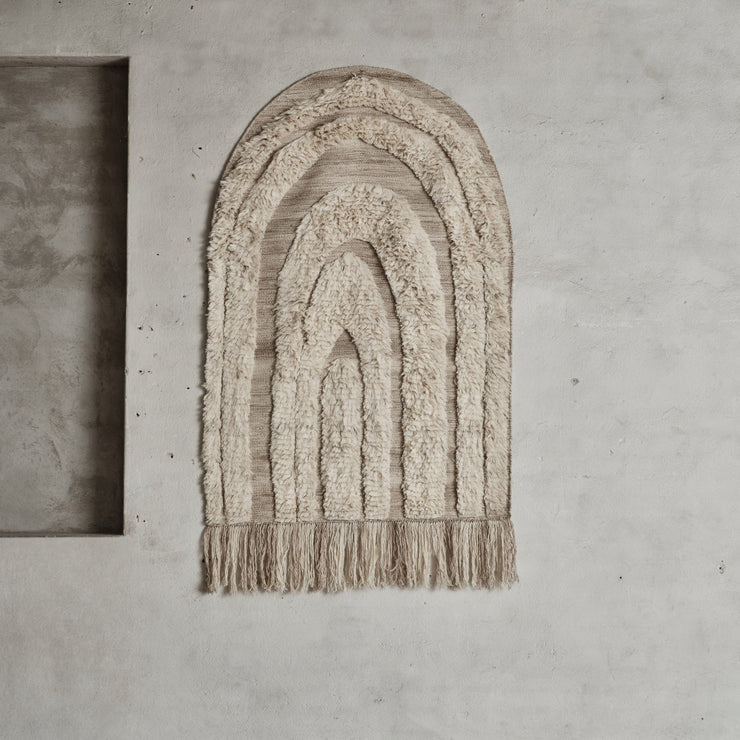 Den buede triumfbuen sammen med variasjonene i lav og høy luvlengde skaper et passe livlig og bohemsk mønster. En lang frynsekant løfter endekanten av teppet og tilfører en fin kunstnerisk detalj. Den flatvevde delen er sandfarget, mens den høyere luven har en myk, off-white farge. Et vakkert og litt spennende teppe som gjør seg veldig fint i en gang, i en stue og på et soverom – enten på gulvet eller på veggen!