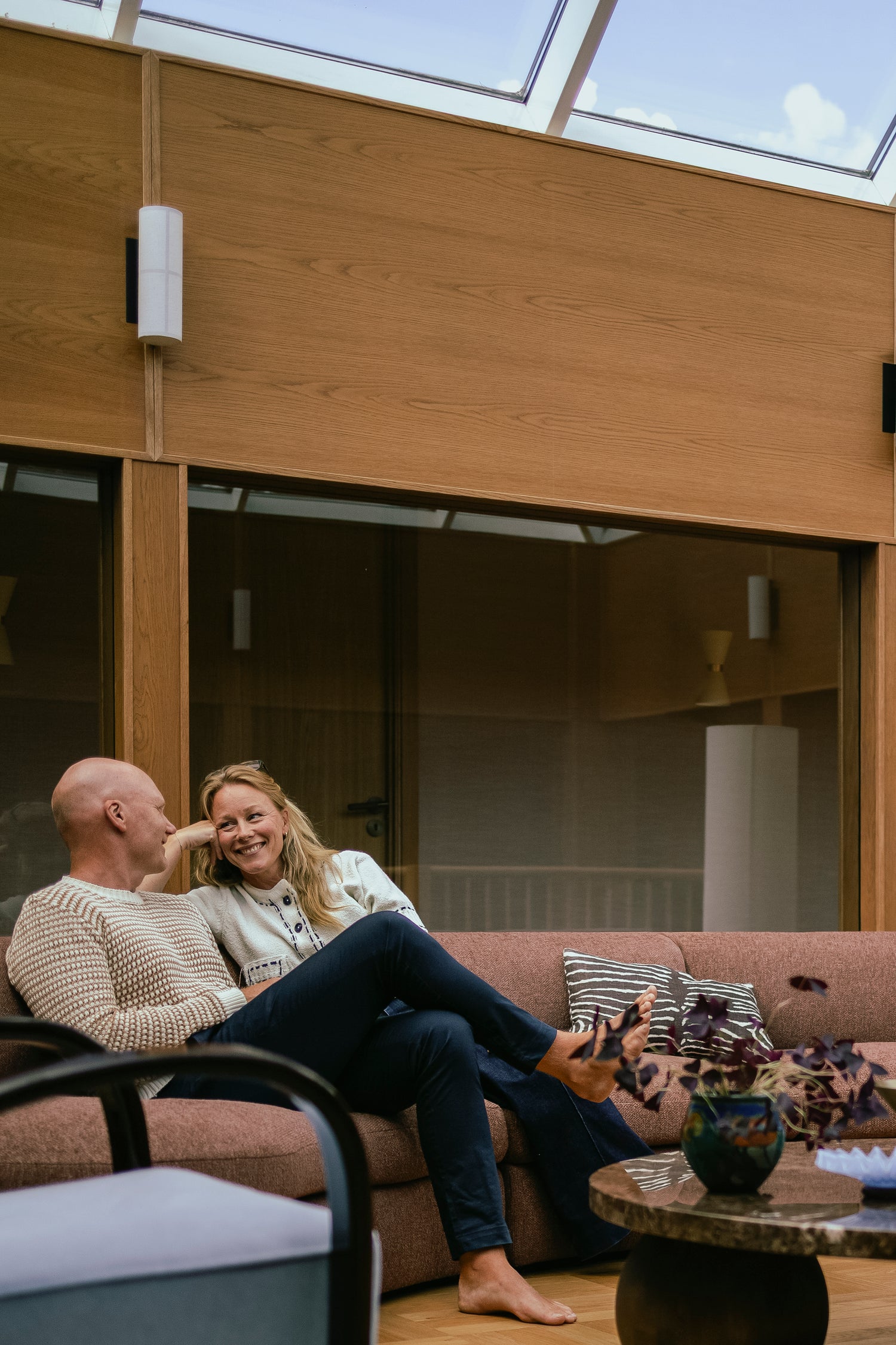 Buet sofa Coda fra New Works i rustfarget bouclétekstil fra OsloDeco i atriumstue renovert av AlexArk. Eierne sitter i sofaen, omringet av finerkledde vegger og vakkert interiør i brunt, messing og blått. Foto: OsloDeco