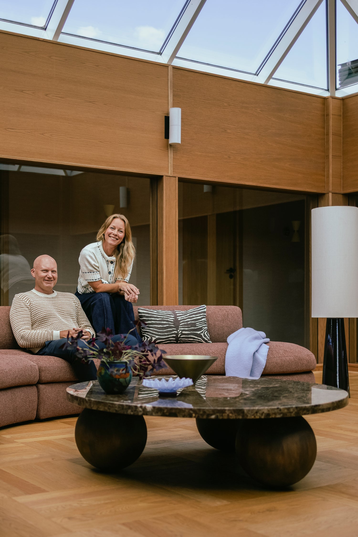 Buet sofa Coda fra New Works i rustfarget bouclétekstil fra OsloDeco i atriumstue renovert av AlexArk. Eierne sitter i sofaen, omringet av finerkledde vegger og vakkert interiør. Foto: OsloDeco