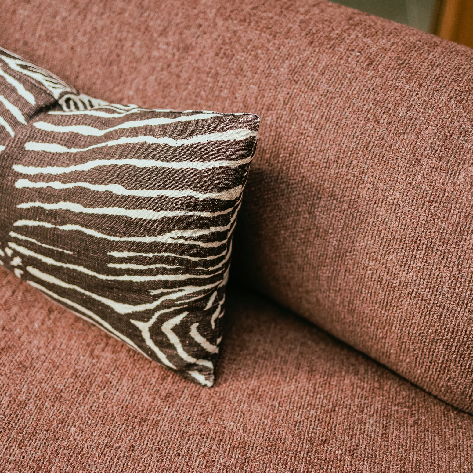 Nærbilde av buet sofa Coda fra OsloDeco. Det myke og slitesterke rustrøde bouclétekstilet sofaen er trukket i er vevd på en måte som gir det et ekstra fint spill. En brun og hvit sebratripet pute komplimenterer. Foto: OsloDeco