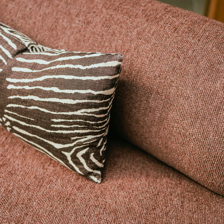 Nærbilde av buet sofa Coda fra OsloDeco. Det myke og slitesterke rustrøde bouclétekstilet sofaen er trukket i er vevd på en måte som gir det et ekstra fint spill. En brun og hvit sebratripet pute komplimenterer. Foto: OsloDeco