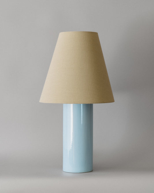 Bordlampen Giro Baby Blue & Beige har en keramisk sylinderformet lampefot med en lyseblå glasur og en konisk skjerm drapert i en teksturert lys beige bomull (fra Haider Ackermann) som sprer et behagelig og stemningsfullt varmt lys. En raffinert bordlampe som vil tilføre ethvert rom en dose sofistikert eleganse!
