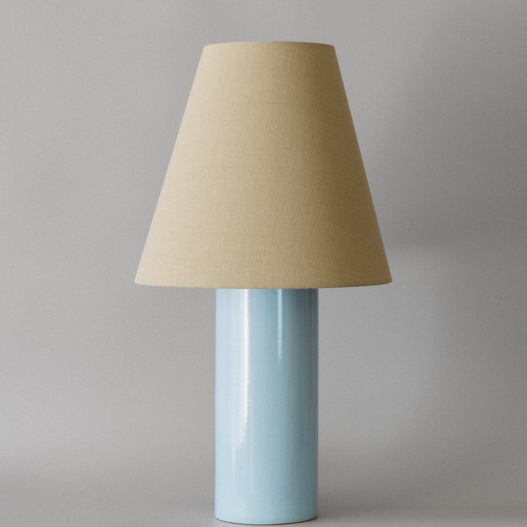 Bordlampen Giro Baby Blue & Beige har en keramisk sylinderformet lampefot med en lyseblå glasur og en konisk skjerm drapert i en teksturert lys beige bomull (fra Haider Ackermann) som sprer et behagelig og stemningsfullt varmt lys. En raffinert bordlampe som vil tilføre ethvert rom en dose sofistikert eleganse!