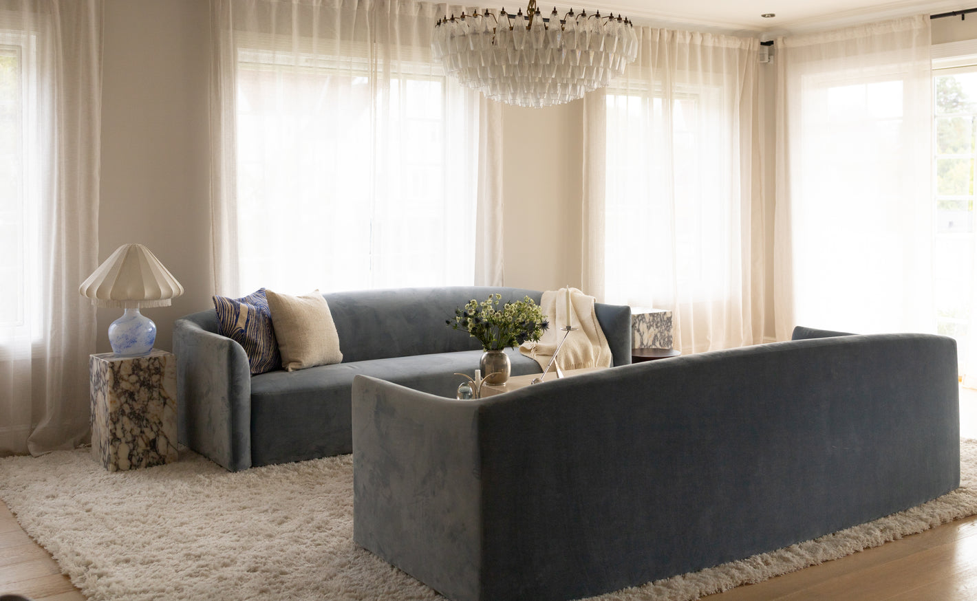 Klassisk og elegant stue med to 3-setere i blått stående mot hverandre, lyse vegger og lett gjennomskinnelige gardiner.  Murano-taklampe og detaljer i mørkt marmor, glass og blank krom. Lyse eikegulv og lyst ullteppe med frynser.