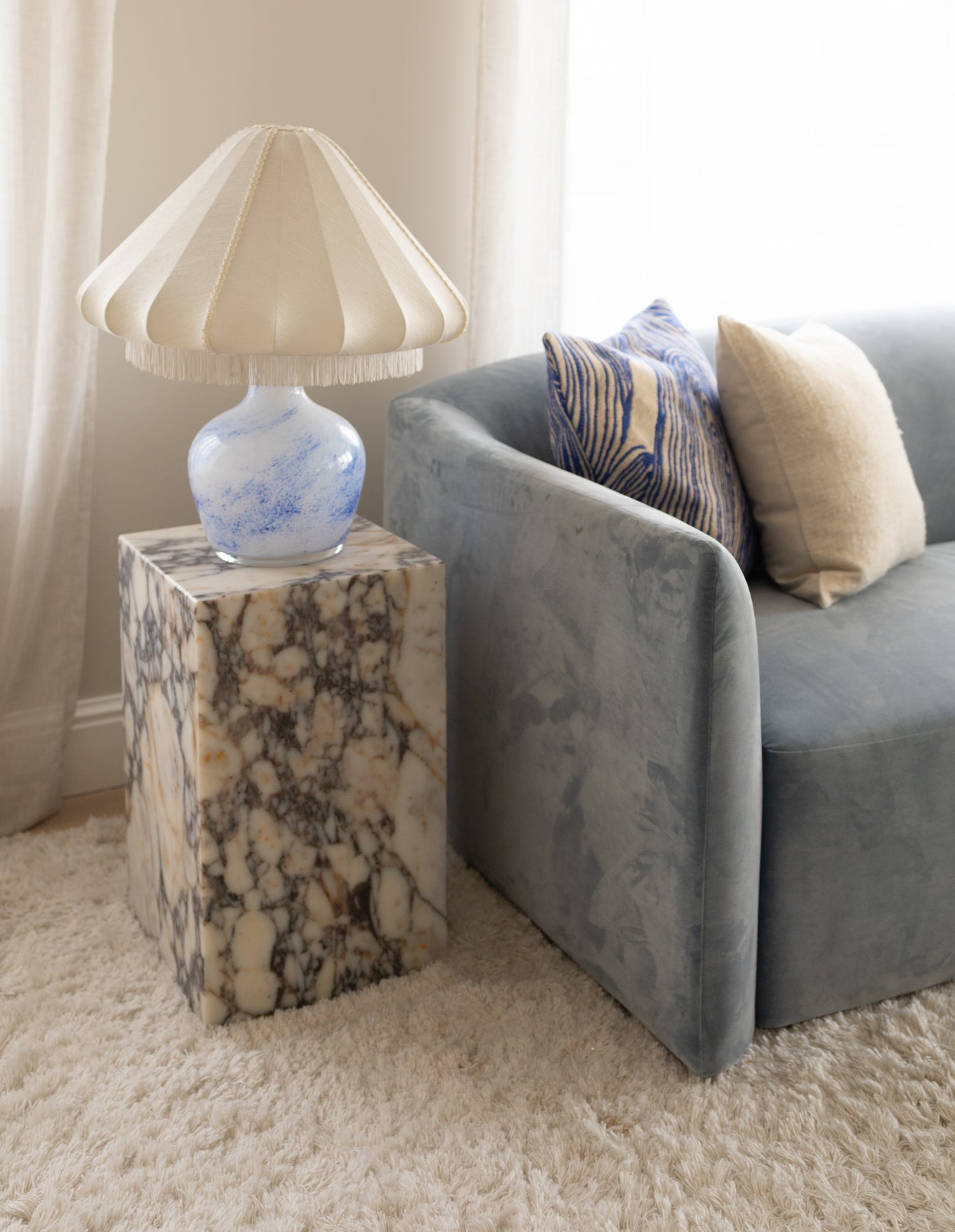 Vintage bordlampe i glass med tekstilskjerm stående på et sidebord i vakker marmor. Dueblå sofa i velur med puter i blått og krem. Lyst ullteppe med frynser.