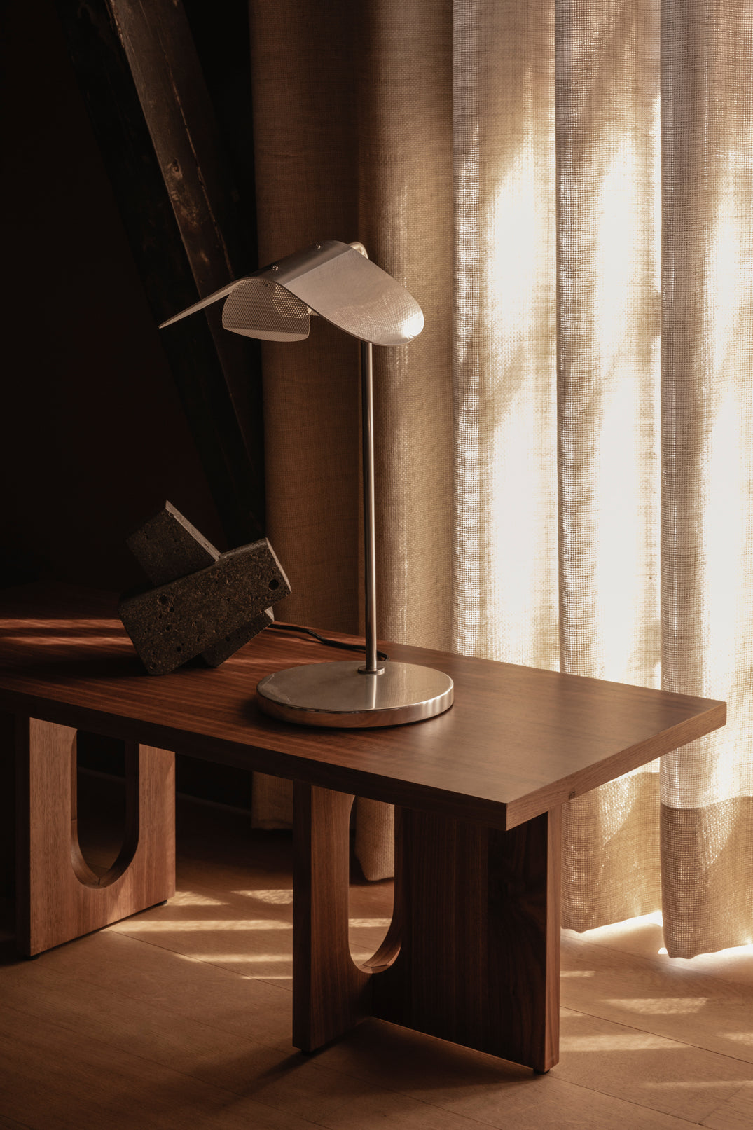Bilde av sofabordet Androgyne Lounge Table fra Menu i valnøtt mot eikegulv og lyse lingardiner. På bordet står en lampe i stål.
