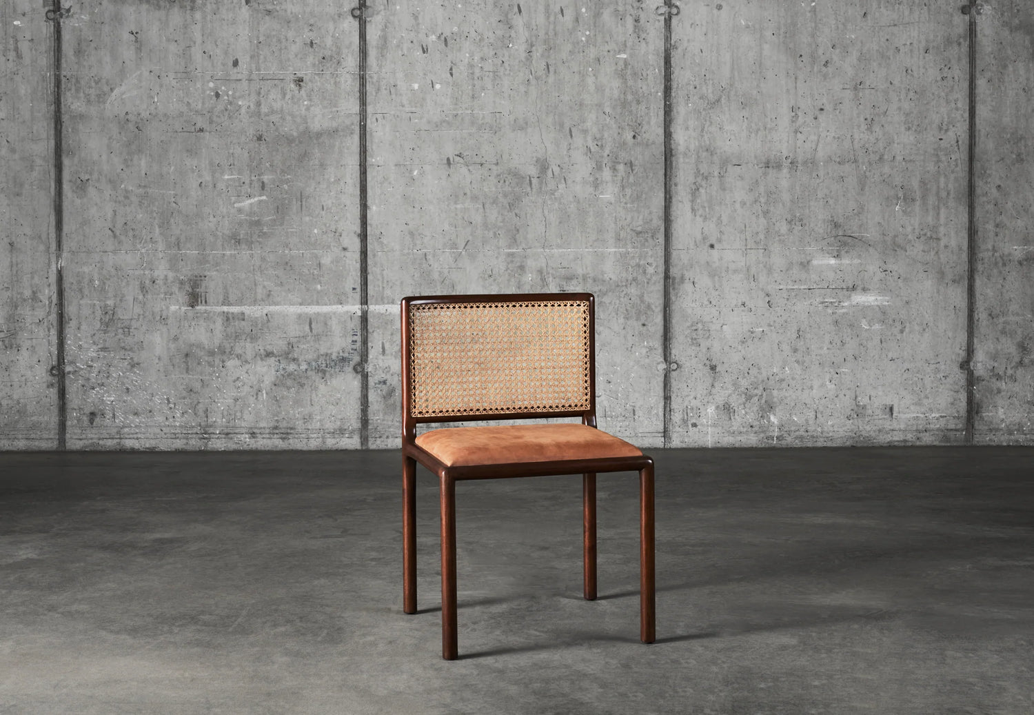Bilde av stolen Mesh Chair fra Dusty Deco i mørk mahogni, rygg i flettet rotting og sete i brunt skinn mot rå betongvegg.