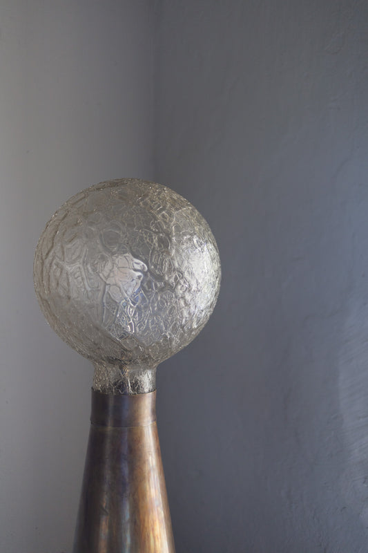 Kombinasjonen av materialene, formen og lyset den avgir er nesten magisk fin: På den store koniske lampefoten i patinert messing hviler en vakker glasskuppel, som med sine unike fasetteringer i glasset reflekterer lyset på nydelig vis når den er tent.
