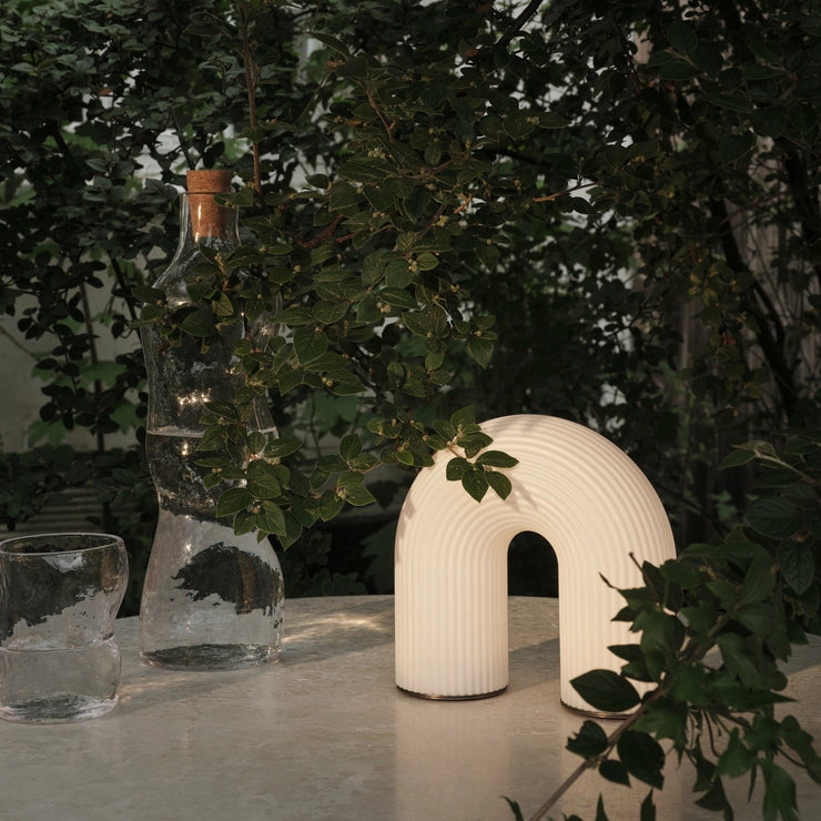 Den nette oppladbare Vuelta-lampen fra Ferm Living er en unik lyskilde, formet som en skulpturell bue som minner om gamle søyler og arkitektur. Den er laget av hvitt opalglass, med en rillet overflate som gir den et appellerende uttrykk. LED-lyskilden gir et mykt, diffust lys, og lampen kan dimmes i fire trinn slik at du kan justere lyset etter behov - og som gjør den til et godt bidrag til bedre stemning i rommet.