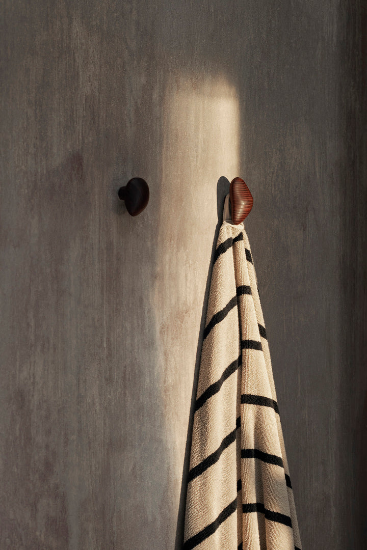 Bilde fra baderom med to mørkebrune knagger i tre fra Ferm Living. På knaggene Cairn er det hengt opp et stripete badehåndkle.